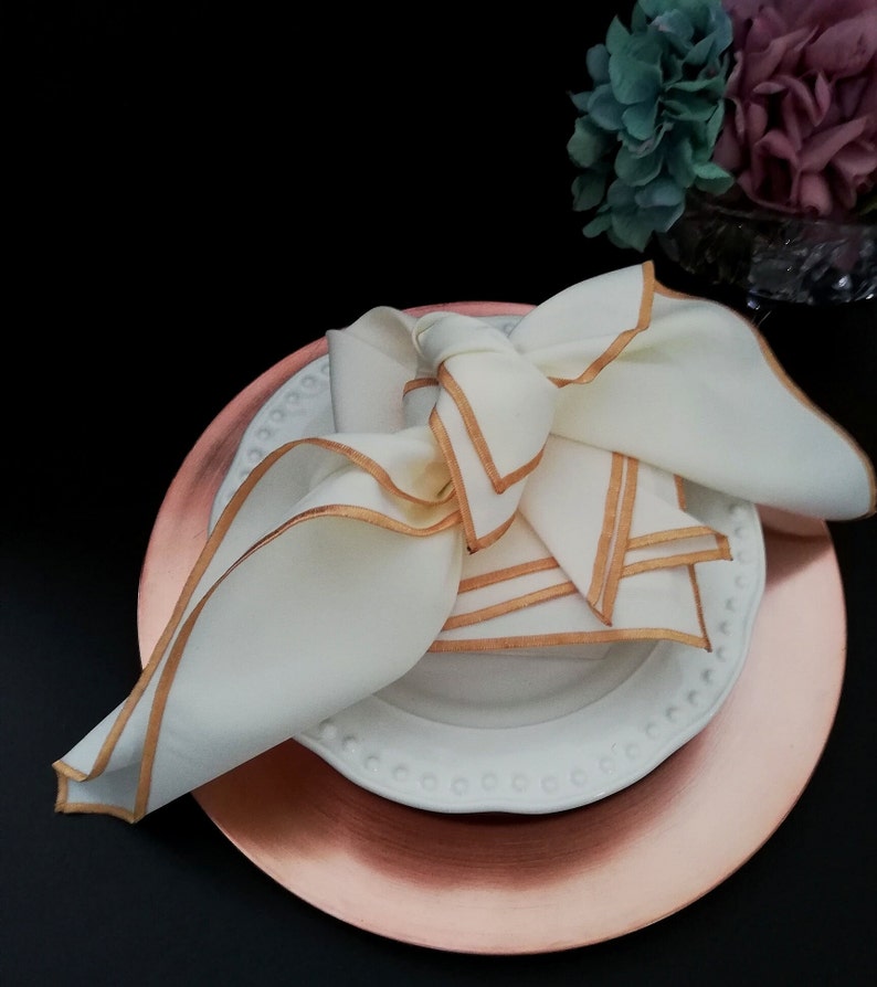 May include: A folded white linen napkin with gold trim sits on a white plate with a beaded edge, placed on a copper-coloured charger plate. The napkin is tied in a decorative knot. The setting is on a black surface.