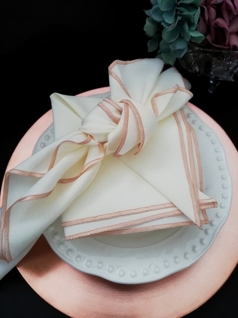 May include: An ivory napkin with rose gold trim, knotted on a white plate with a beaded edge, atop a rose gold charger plate. A stylish table setting for a special occasion.