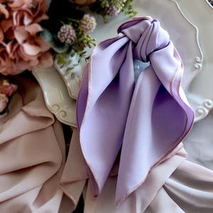 Pu&ograve; includere: Una sciarpa di seta color lavanda annodata, con un bordo rosa chiaro a contrasto. La sciarpa &egrave; drappeggiata su un piatto decorativo bianco, con uno sfondo sfocato di fiori e altri tessuti. La consistenza morbida e la tavolozza dei colori creano un'estetica delicata ed elegante.
