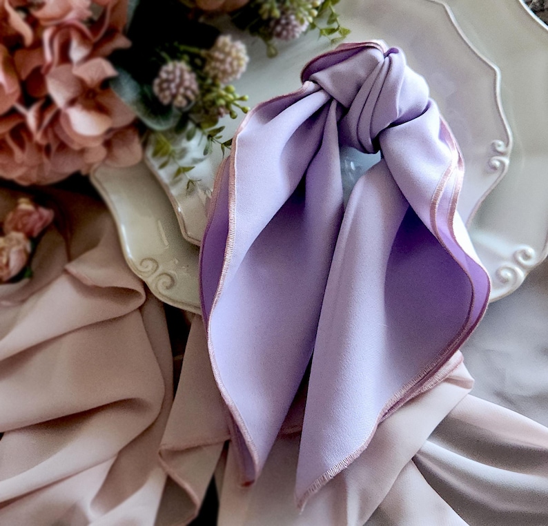 Pu&ograve; includere: Una sciarpa di seta color lavanda annodata, con un bordo rosa chiaro a contrasto. La sciarpa &egrave; drappeggiata su un piatto decorativo bianco, con uno sfondo sfocato di fiori e altri tessuti. La consistenza morbida e la tavolozza dei colori creano un'estetica delicata ed elegante.