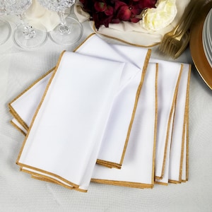 May include: Several white cloth napkins with gold edging are arranged on a white tablecloth. The image also shows crystal glassware, gold cutlery, and a gold-rimmed plate, suggesting a formal dining setting. The napkins are neatly folded.