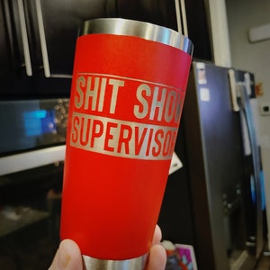 May include: Red stainless steel tumbler with a silver band and the text "Shit Show Supervisor" engraved on the band.