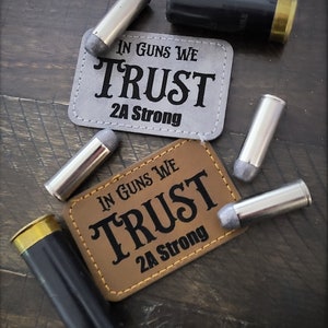 May include: An overhead shot features two rectangular patches with the text "IN GUNS WE TRUST 2A Strong" in black. Several silver and black shotgun shells and bullets are scattered around the patches on a dark wooden surface.