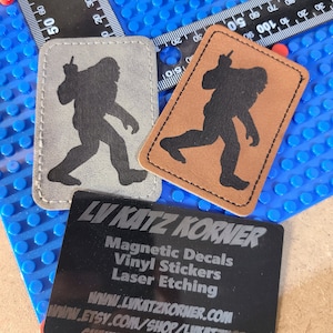 May include: Two leather patches with a black silhouette of a Bigfoot figure. The patches are on a blue background with a black business card that reads "LV KATZ KORNER Magnetic Decals Vinyl Stickers Laser Etching www.LVKATZKORNER.COM www.ETSY.COM/SHOP/LUKATZKORNER SHAUN.BULV@GMAIL.COM"