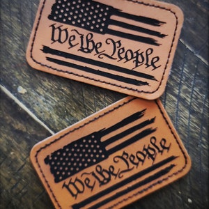 May include: Two brown leather patches with a black distressed American flag and the text "We the People" in a black script font.