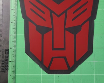 May include: A red and black Autobot logo sticker. The sticker is a shield-shaped emblem with a red face and black outlines. The sticker is approximately 5 inches tall and 4 inches wide.