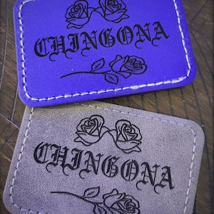 May include: Two leather patches with a stitched border. The top patch is purple with the word "CHINGONA" in black script and two roses. The bottom patch is gray with the same word and roses.