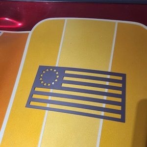 May include: A dark gray metal cutout of the American flag with a circle of stars in the upper left corner and horizontal stripes. The flag is set against a yellow and orange background.