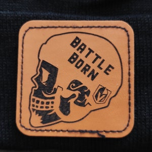 May include: A brown leather patch with a black skull wearing a helmet that says "BATTLE BORN" on it. The skull has a black and white design on the helmet.