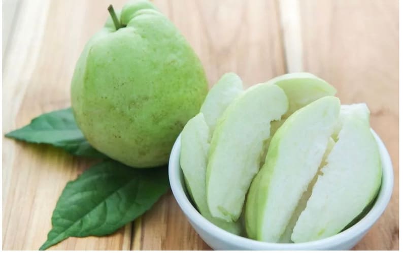 May include: A whole green guava and several slices of guava in a white bowl on a wooden surface.