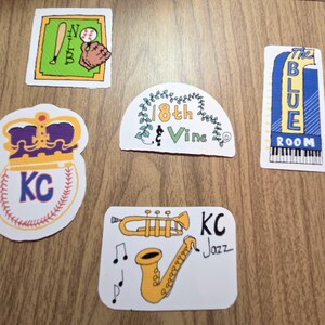 May include: A collection of colorful stickers on a wooden surface. The stickers feature baseball, jazz instruments, and text including "18th Vine," "The Blue Room," and "KC Jazz." The baseball sticker has a crown and the letters "KC."