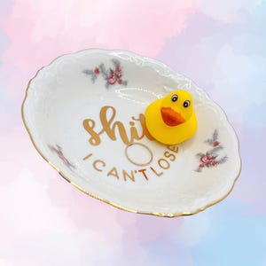 Könnte beinhalten: Eine weiße Keramik-Schmuckschale mit goldenen Verzierungen und floralen Akzenten, mit dem Text "shit I can't lose" in goldfarbenen Buchstaben. Eine gelbe Quietscheente und ein goldener Ring befinden sich in der Schale.