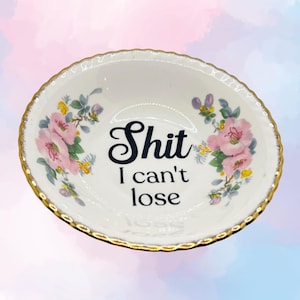 Könnte beinhalten: Eine weiße Keramikschale mit goldenem Rand, mit rosa Blumendesigns und dem Text "Shit I can't lose" in schwarzer Schrift. Die Schale hat einen gewellten Rand und wird wahrscheinlich zu Dekorationszwecken verwendet.
