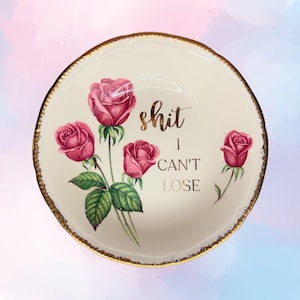 May include: A white ceramic trinket dish with a gold rim and a floral design. The dish has the text "shit I can't lose" in gold lettering.
