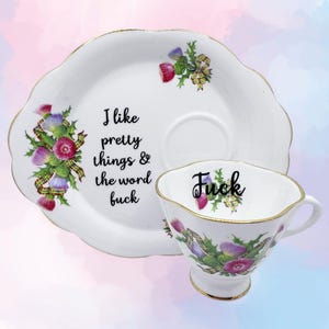 Könnte beinhalten: Ein weißes Porzellan-Tassenset mit Untertasse und goldfarbenen, gewellten Rändern. Die Untertasse zeigt ein Distel-Blumenmuster und den Text, I like pretty things & the word fuck. Die Tasse zeigt ein Distel-Design und das Wort Fuck.