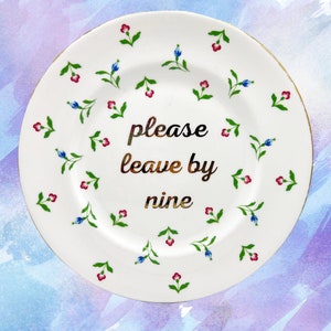 Peut inclure: Une assiette en céramique blanche avec un bord doré et un motif floral. L'assiette porte l'inscription "please leave by nine" en lettres dorées.