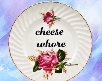 May include: White ceramic plate with a gold rim and a floral design featuring pink roses and green leaves. The plate has the words "cheese whore" in black script. The background is a watercolor wash of blue and purple.