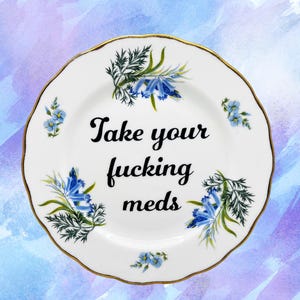 Op de afbeelding: Witte keramische plaat met een gouden rand, versierd met blauwe bloemmotieven en de tekst "Take your fucking meds" in zwart schrift. De achtergrond is een aquarel wassing in blauw en paars.