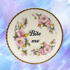 May include: A white ceramic saucer with a gold rim, featuring pink floral designs and the words "Bite me" in black script. The saucer has a vintage aesthetic, with a delicate and ornate appearance.