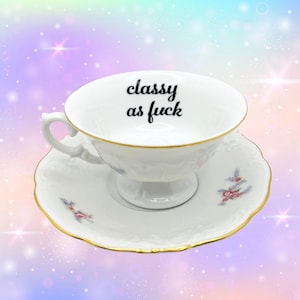 Peut inclure: Une tasse à thé et sa soucoupe en porcelaine blanche avec un liseré doré et des motifs floraux délicats. L'intérieur de la tasse présente le texte noir "classy as fuck" en écriture cursive. L'ensemble possède un bord festonné et un pied.