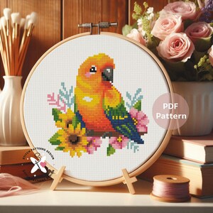 May include: A cross-stitch pattern of a colorful parrot with a yellow and orange body, black beak, and blue wings, surrounded by flowers. The design is in a wooden embroidery hoop, with the text "PDF Pattern" visible.