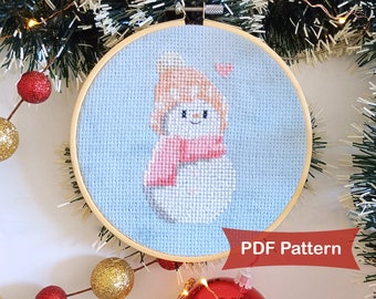 May include: A cross-stitch pattern of a snowman wearing a pink scarf and a peach-colored hat. The snowman is on a light blue background with a small pink heart above it. The pattern is in a wooden embroidery hoop. The text "PDF Pattern" is in red at the bottom of the image.