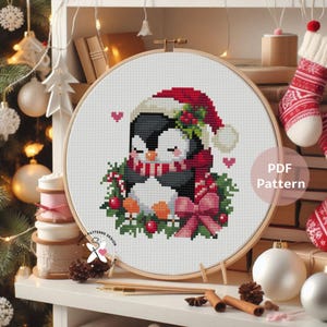 May include: A cross-stitch pattern of two penguins hugging, wearing a Santa hat and scarf, surrounded by a wreath and a pink bow. The design is in a wooden embroidery hoop, with the text "PDF Pattern" visible.