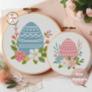 Puede incluir: Dos patrones de punto de cruz en aros de madera. Uno presenta un diseño de huevo de Pascua azul con detalles florales, el otro un huevo rosa con elementos florales similares. Los patrones están rodeados de flores rosas y blancas.