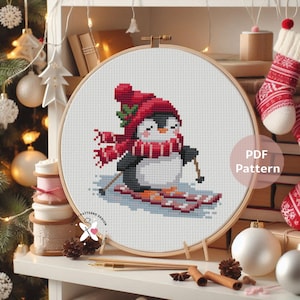 May include: A cross-stitch pattern of a penguin skiing, wearing a red hat and scarf. The penguin is on skis, with a wooden embroidery hoop. The text "PDF Pattern" is visible.