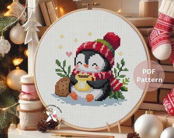 May include: A cross-stitch pattern featuring a cute penguin wearing a red and white hat and scarf, holding a mug. The penguin is surrounded by green sprigs and a cookie. The design is set in a wooden embroidery hoop. The words "PDF Pattern" are visible.