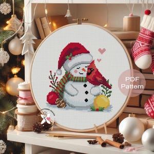 May include: A cross-stitch pattern in a wooden hoop features a snowman wearing a red hat and green striped scarf, embracing a red cardinal. The design includes a red apple, a yellow ornament, and the text "PDF Pattern".