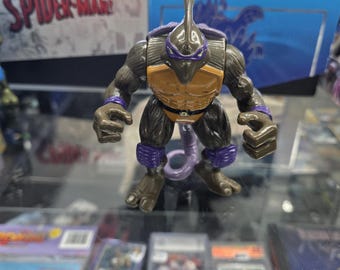 May include: A detailed action figure of a humanoid insect with a brown and tan body, purple accents, and a long, curved horn. The figure is posed on a glass surface, with trading cards and boxes in the background.