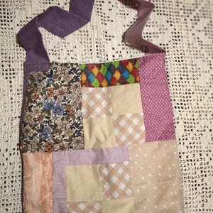Peut inclure: Un sac fourre-tout patchwork avec une sangle violette. Le sac présente divers motifs de tissu, notamment des motifs floraux, à pois, à carreaux et étoilés. La palette de couleurs comprend des nuances de rose, de beige et de violet.