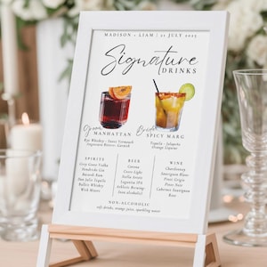 Pode incluir: Um cartaz branco com um cavalete de madeira, listando as bebidas de assinatura para um casamento. O cartaz tem um título escrito à mão "Signature Drinks" e lista dois coquetéis, "Manhattan" e "Spicy Marg", com seus ingredientes. O cartaz também lista as opções de bebidas alcoólicas, cerveja e vinho.