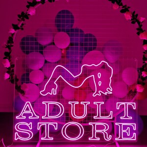 Peut inclure: Enseigne au n&eacute;on avec un fond rose et un contour blanc. L'enseigne indique "ADULT STORE" et pr&eacute;sente une silhouette stylis&eacute;e d'une femme.
