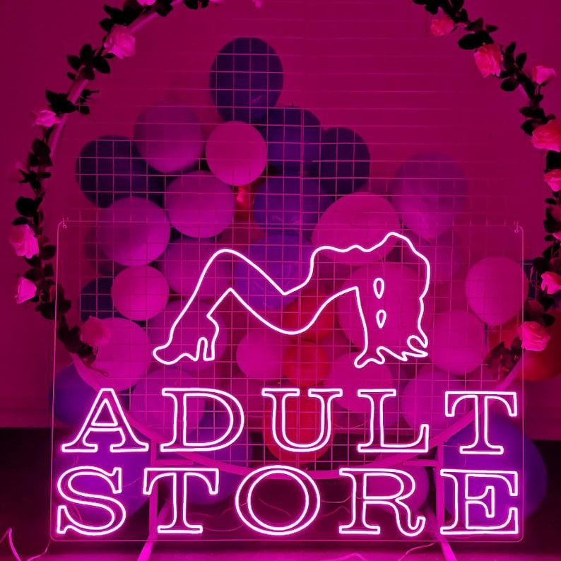 Peut inclure: Enseigne au n&eacute;on avec un fond rose et un contour blanc. L'enseigne indique "ADULT STORE" et pr&eacute;sente une silhouette stylis&eacute;e d'une femme.