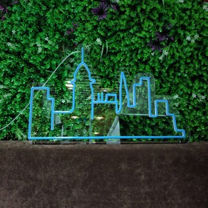 May include: A blue neon sign depicting a city skyline with tall buildings and a spire. The sign is mounted on a dark brown surface in front of a green wall.