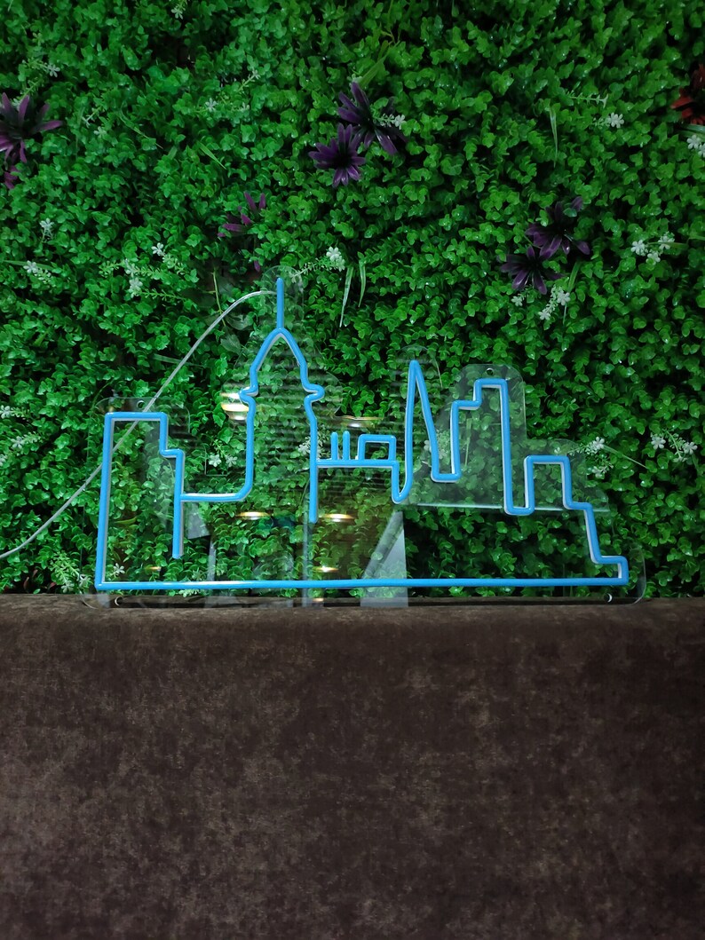 May include: A blue neon sign depicting a city skyline with tall buildings and a spire. The sign is mounted on a dark brown surface in front of a green wall.