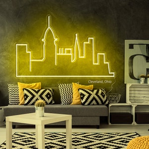 May include: A neon sign depicting the Cleveland, Ohio skyline in a yellow glow. The sign is mounted on a dark gray wall in a living room setting. The sign is surrounded by a gray couch with black and white patterned pillows and a yellow throw pillow.