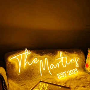 Peut inclure: Un panneau lumineux blanc en n&eacute;on avec le texte "The Martins EST 2021" sur une surface blanche et moelleuse. Le panneau est &eacute;clair&eacute; d'une lueur jaune chaude.