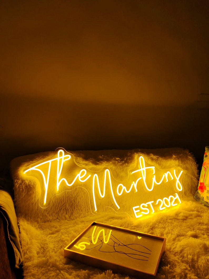 Peut inclure: Un panneau lumineux blanc en n&eacute;on avec le texte "The Martins EST 2021" sur une surface blanche et moelleuse. Le panneau est &eacute;clair&eacute; d'une lueur jaune chaude.