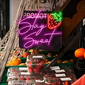 Peut inclure: Un panneau n&eacute;on rose avec le texte "Stay Sweet" et un graphique de fraise rouge et verte. Le panneau est suspendu au-dessus d'un pr&eacute;sentoir de fraises fra&icirc;ches dans des contenants en plastique transparents.