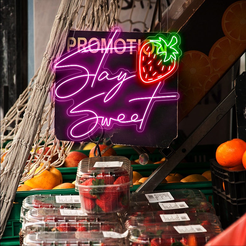Peut inclure: Un panneau n&eacute;on rose avec le texte "Stay Sweet" et un graphique de fraise rouge et verte. Le panneau est suspendu au-dessus d'un pr&eacute;sentoir de fraises fra&icirc;ches dans des contenants en plastique transparents.