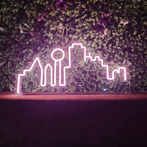 Pu&ograve; includere: Un'insegna al neon rosa a forma di skyline cittadino, posta su uno sfondo di fogliame verde e fiori viola. Il contorno luminoso dell'insegna crea un contrasto sorprendente con lo sfondo naturale. Design moderno per l'arredamento d'interni.