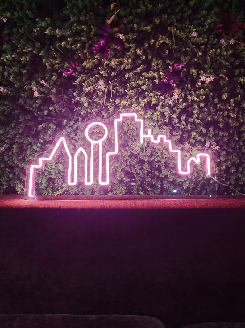 Pu&ograve; includere: Un'insegna al neon rosa a forma di skyline cittadino, posta su uno sfondo di fogliame verde e fiori viola. Il contorno luminoso dell'insegna crea un contrasto sorprendente con lo sfondo naturale. Design moderno per l'arredamento d'interni.