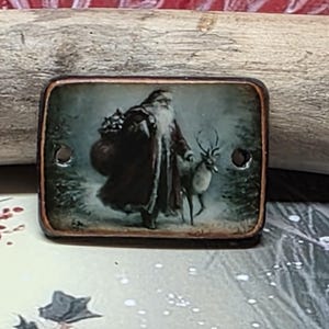May include: A rectangular pendant featuring a vintage-style illustration of Santa Claus in a long red coat carrying a sack, accompanied by a reindeer in a snowy forest setting. The piece has a dark brown border and two side holes for threading.