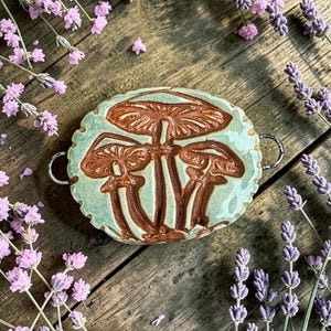 May include: A round, light green ceramic pendant with a scalloped edge featuring a relief design of three brown mushrooms. The piece has a metallic loop on each side for attachment, showcasing a glossy finish and detailed texture on the mushroom caps.