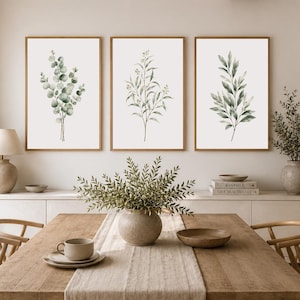May include: A set of three framed wall art prints featuring botanical illustrations of green eucalyptus and leafy branches on a white background, displayed in light wood frames above a wooden dining table with a beige table runner.