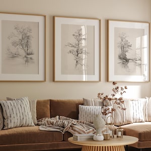 Puede incluir: Un sofá seccional marrón con una mesa de café de madera y tres dibujos en blanco y negro enmarcados de árboles en la pared detrás de él. El sofá tiene una manta de lana marrón claro y varias almohadas.