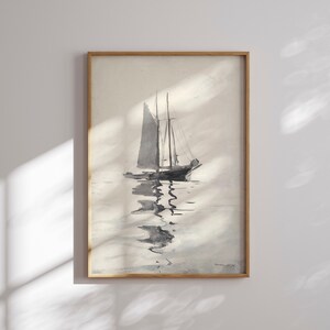 Op de afbeelding: Een ingelijste aquarel van een zeilboot op kalm water. Het kunstwerk is voornamelijk in grijstinten en zwart, met de boot en zijn weerspiegeling duidelijk zichtbaar. De lijst is gemaakt van lichtgekleurd hout, wat een klassieke touch geeft aan het nautische thema.